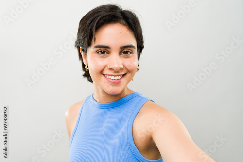 Smiling young woman with short hair in a blue top taking a selfie against a white background