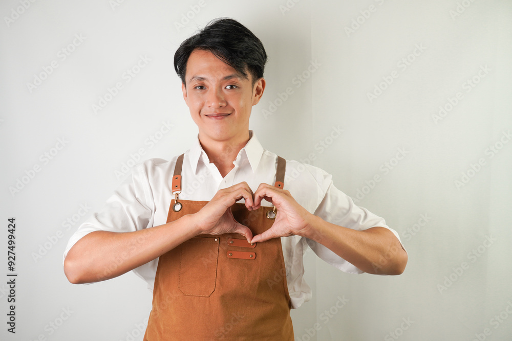Friendly young asian man wearing rolled-up white sleeve shirt and brown apron with love heart shaped finger hand gesture. Isolated over white background.