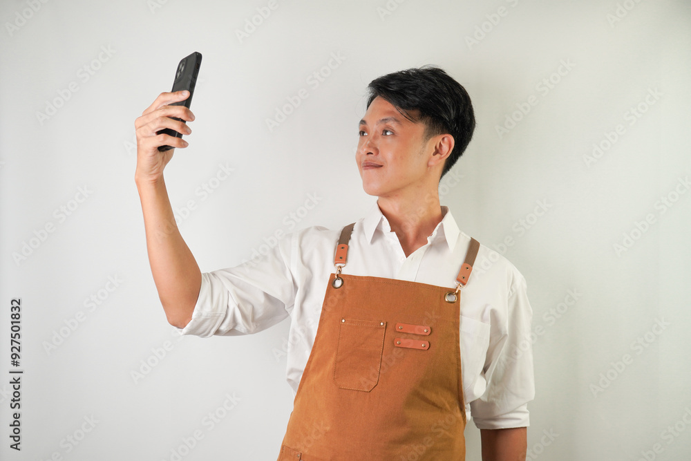 Confident young asian man wearing rolled-up white sleeve shirt and brown apron is holding a smartphone with smiling face expression. Isolated over white background. Confident young asian man wearing rolled-up white sleeve shirt and brown apron is holding a smartphone with smiling face expression. Isolated over white background.