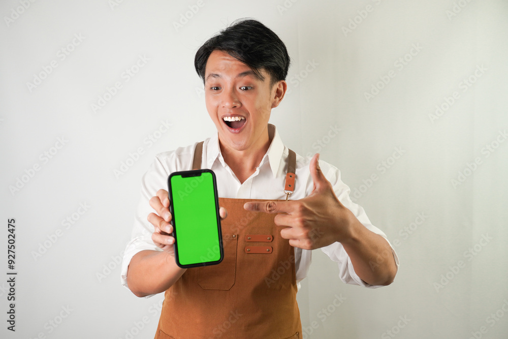 Excited young asian man wearing rolled-up white sleeve shirt and brown apron is pointing vertical blank green screen smartphone with smiling face expression. Isolated over white background. Excited young asian man wearing rolled-up white sleeve shirt and brown apron is pointing vertical blank green screen smartphone with smiling face expression. Isolated over white background.