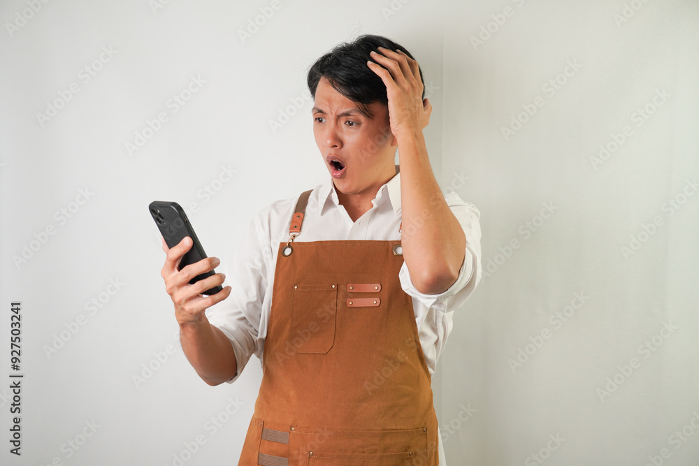 Shocked young asian man wearing rolled-up white sleeve shirt and brown apron is holding a smartphone with surprised face expression. Isolated over white background. Shocked young asian man wearing rolled-up white sleeve shirt and brown apron is holding a smartphone with surprised face expression. Isolated over white background.