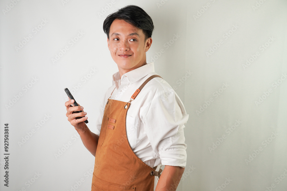 Confident young asian man wearing rolled-up white sleeve shirt and brown apron is holding a smartphone with smiling face expression. Isolated over white background. Confident young asian man wearing rolled-up white sleeve shirt and brown apron is holding a smartphone with smiling face expression. Isolated over white background.