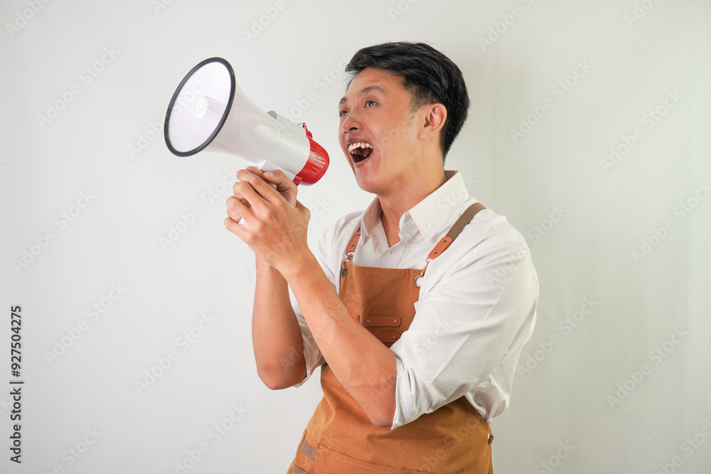 Excited young asian man wearing rolled-up white sleeve shirt and brown apron is holding using megaphone with smiling face expression. Screaming, shouting at loudspeaker. Isolated over white background Excited young asian man wearing rolled-up white sleeve shirt and brown apron is holding using megaphone with smiling face expression. Screaming, shouting at loudspeaker. Isolated over white background