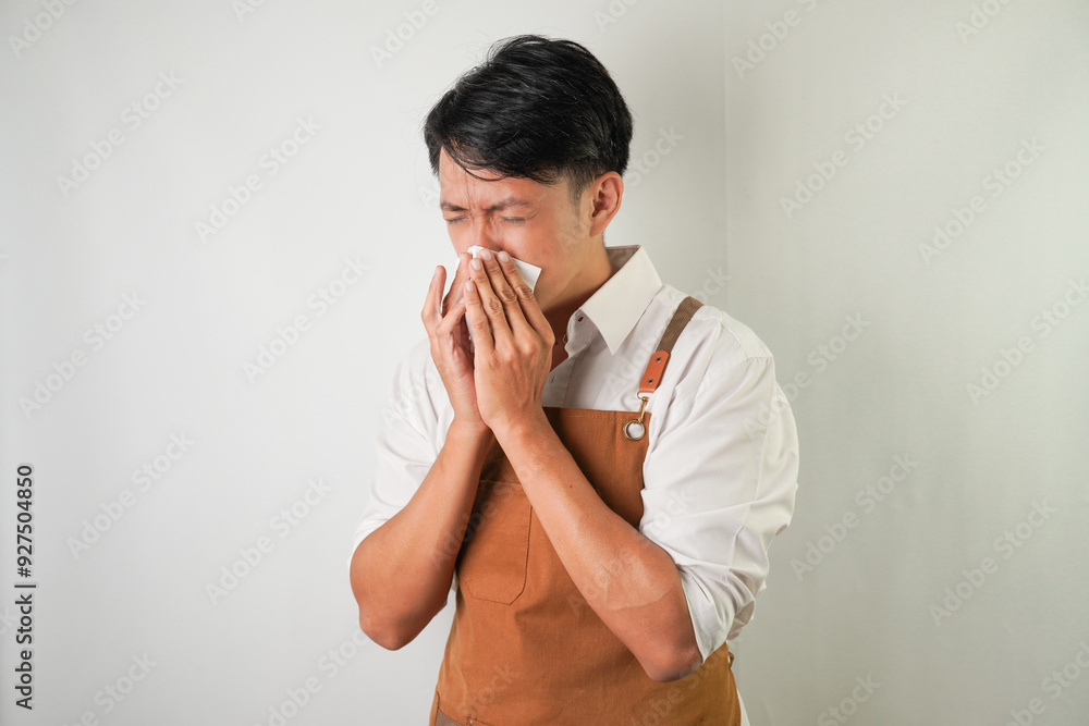 Young asian man wearing rolled-up white sleeve shirt and brown apron is sneezing and holding tissue paper with grimacing face expression. Isolated over white background. Young asian man wearing rolled-up white sleeve shirt and brown apron is sneezing and holding tissue paper with grimacing face expression. Isolated over white background.