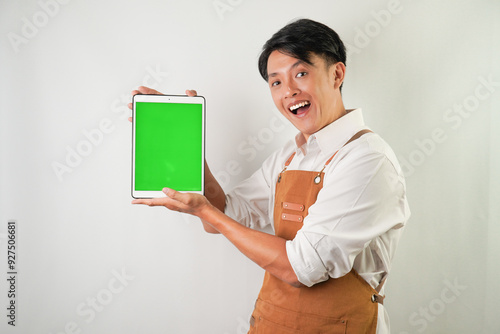 Surprised amazed young asian man wearing rolled-up white sleeve shirt and brown apron is showing vertical blank green screen tablet with smiling face expression. Isolated over white background.