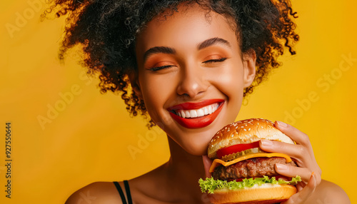 A woman with curly hair is holding a hamburger and smiling. Concept of happiness and enjoyment, as the woman is posing with her food and he is in a good mood