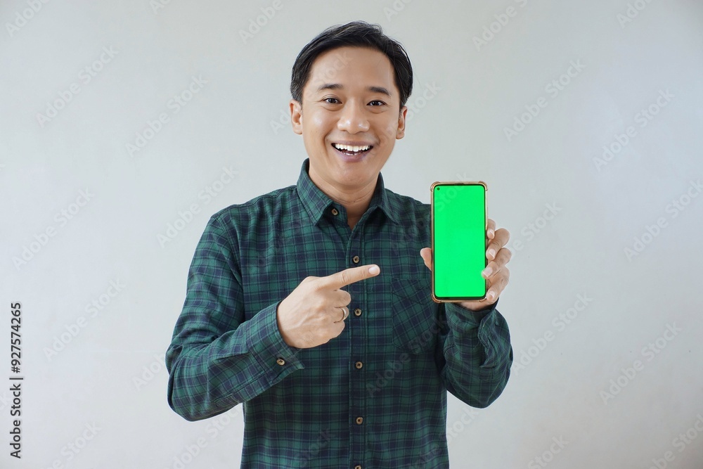 Adult Asian man in green long sleeve flannel shirt showing smartphone display with green screen to camera with left hand while right hand pointing finger to screen. Happy and cheerful expression. Adult Asian man in green long sleeve flannel shirt showing smartphone display with green screen to camera with left hand while right hand pointing finger to screen. Happy and cheerful expression.