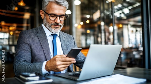 A middle-aged Caucasian man in a suit checks his smartphone while working on a laptop in a modern office setting, surrounded by a stylish, urban atmosphere.