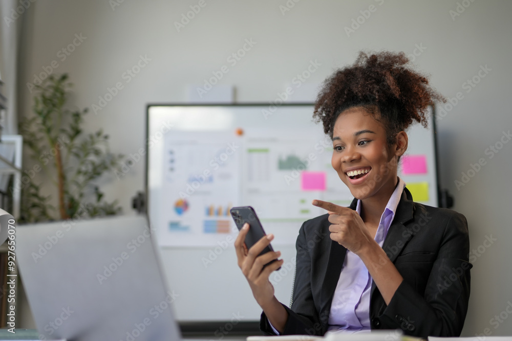 Young businesswoman is smiling and pointing at her smartphone while working in an office with a whiteboard in the background Young businesswoman is smiling and pointing at her smartphone while working in an office with a whiteboard in the background