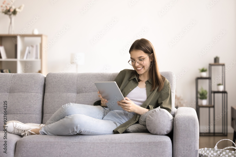 Smiling teenager girl in glasses resting seated on couch with tablet device, chatting on-line, web surfing website, search information. Modern wireless tech usage for fun and leisure. Blogging, gen Z Smiling teenager girl in glasses resting seated on couch with tablet device, chatting on-line, web surfing website, search information. Modern wireless tech usage for fun and leisure. Blogging, gen Z