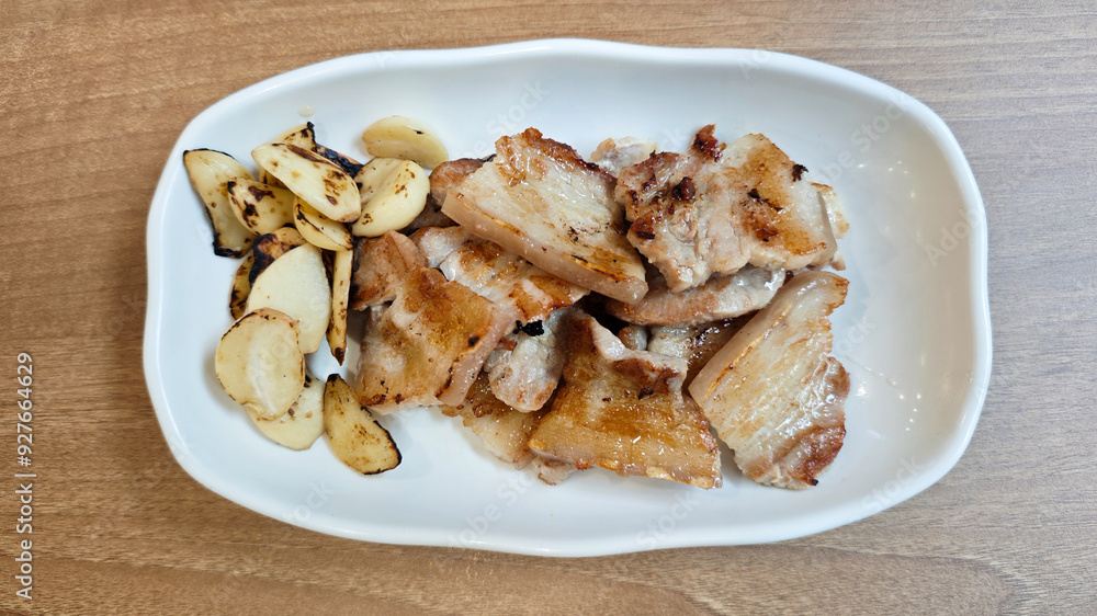 Korean Pork Belly Barbeque Grilled with Roasted Garlic on a White Plate with Light Wooden Table Background Korean Pork Belly Barbeque Grilled with Roasted Garlic on a White Plate with Light Wooden Table Background
