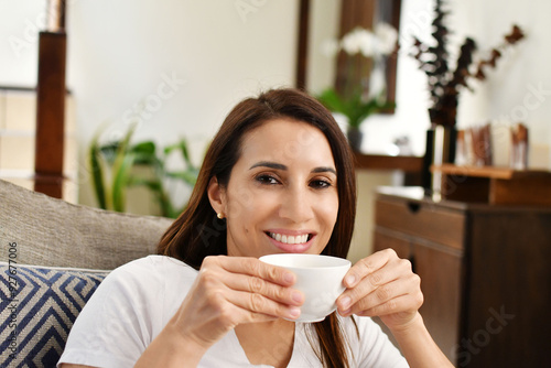 Chica bonita sonriendo a la cámara mientras toma una deliciosa taza de café caliente por la mañana. Toma horizontal.