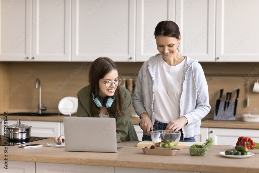 Teen studying in kitchen while her smiling mother prepare lunch for family, lead friendly talk, advising her about essay theme, help with task. Understanding, two generation communication, lifestyle Teen studying in kitchen while her smiling mother prepare lunch for family, lead friendly talk, advising her about essay theme, help with task. Understanding, two generation communication, lifestyle