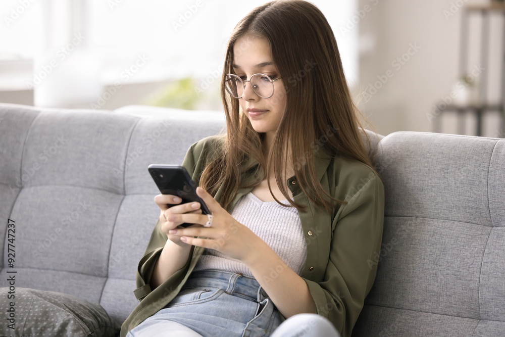 Beautiful teenager girl using modern gadget seated on sofa, wear glasses read messages on smart phone, browsing website, search information, chatting on-line with boyfriend spend leisure on internet Beautiful teenager girl using modern gadget seated on sofa, wear glasses read messages on smart phone, browsing website, search information, chatting on-line with boyfriend spend leisure on internet