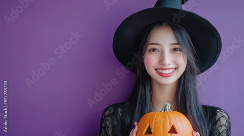 Halloween theme with a young Asian woman in a black costume wearing a witch hat smiling and holding a pumpkin lantern against a purple background