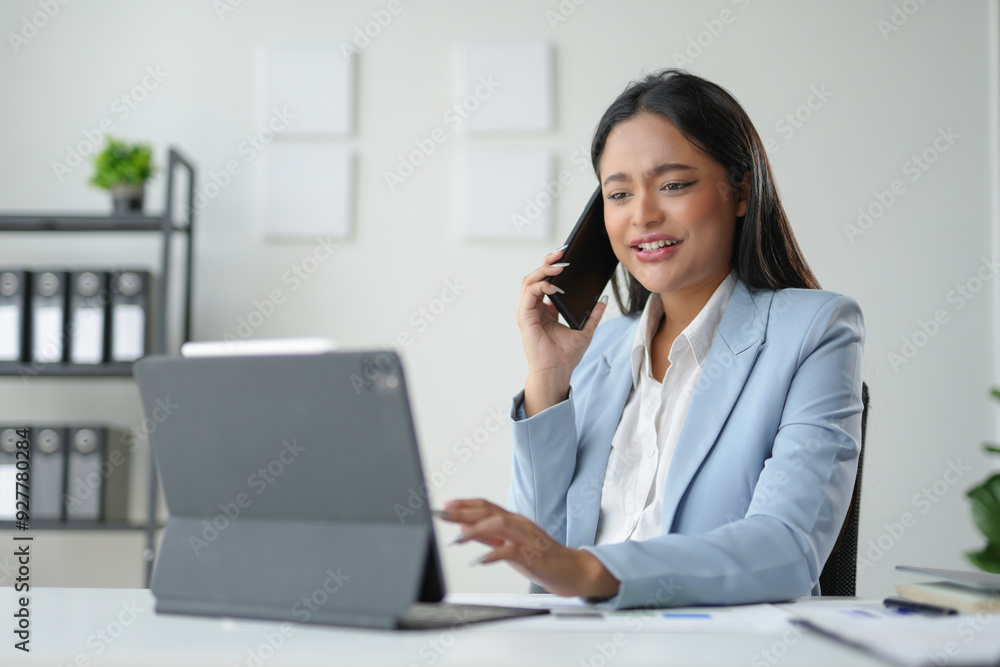 Businesswoman is smiling while talking on the phone and using a digital tablet at her desk in an office Businesswoman is smiling while talking on the phone and using a digital tablet at her desk in an office