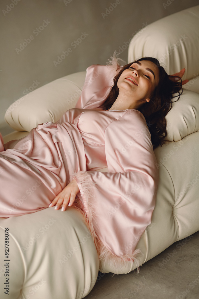 Beautiful female model is lying on a sofa. Portrait of smiling woman In silk pink fashion robe with gorgeous hair style and natural makeup Beautiful female model is lying on a sofa. Portrait of smiling woman In silk pink fashion robe with gorgeous hair style and natural makeup