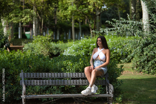 Fotografía Young woman, brunette, beautiful, very tattooed, with white top and jeans, sitting on a park bench, pensive and looking at infinity