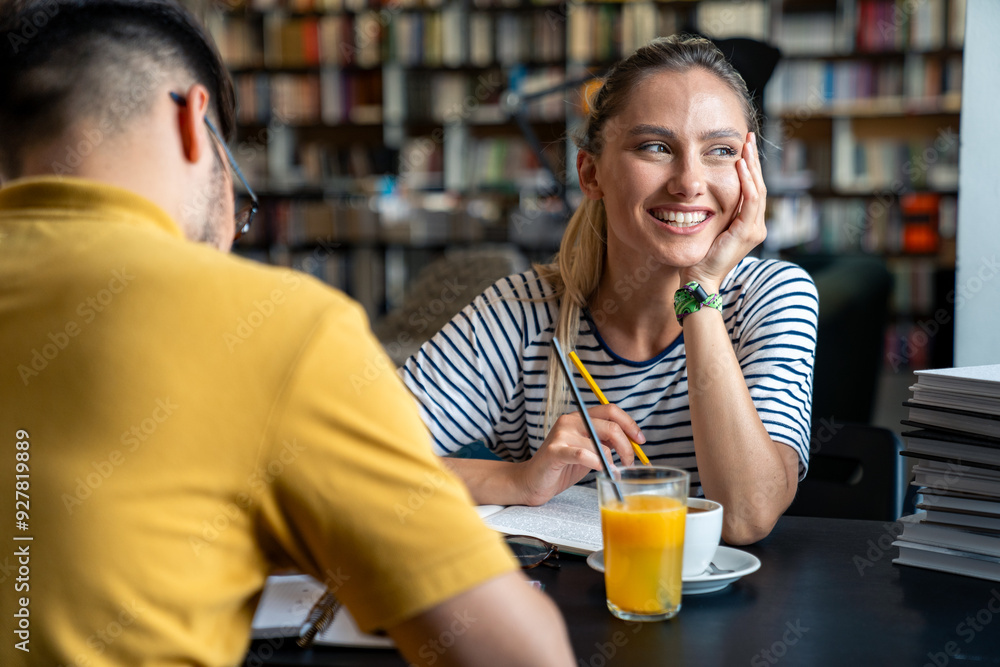 A male and female student sit at a library table surrounded by books, engaging in discussion over drinks, with the female smiling and focused. A male and female student sit at a library table surrounded by books, engaging in discussion over drinks, with the female smiling and focused.