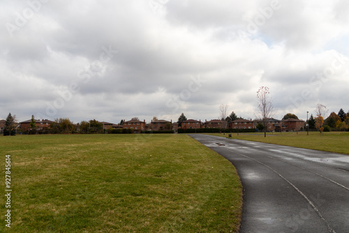 Overcast sky above a winding park path leading towards suburban houses, flanked by expansive green lawns and sparse autumn trees. Taken in Toronto, Canada. photo