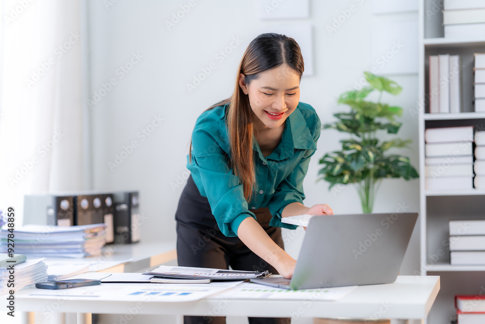 Focused Professional Embraces Success: A young Asian businesswoman beams at her laptop, finding accomplishment and productivity in her modern, light-filled office. Focused Professional Embraces Success: A young Asian businesswoman beams at her laptop, finding accomplishment and productivity in her modern, light-filled office.