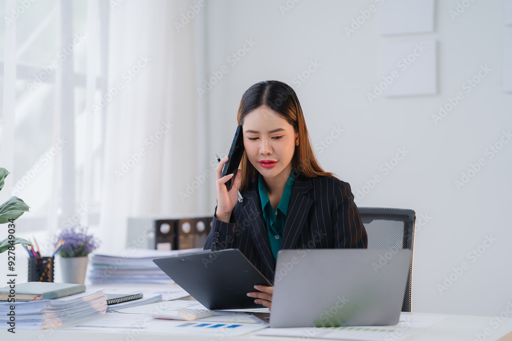 Businesswoman on the Phone: Concentrated and focused, a businesswoman in a professional suit talks on the phone, carefully reviewing documents and data on her laptop, her expression conveys a sense of Businesswoman on the Phone: Concentrated and focused, a businesswoman in a professional suit talks on the phone, carefully reviewing documents and data on her laptop, her expression conveys a sense of