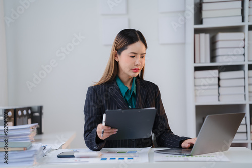 Focused Professional: A dedicated businesswoman meticulously analyzes data on her laptop, displaying a determined focus and commitment to success. Her workspace, cluttered with documents and a smartph photo