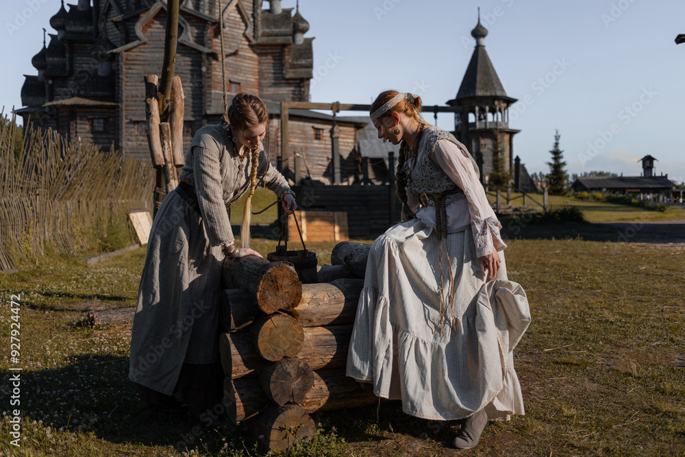 Fototapeta premium young girls in folk costume take water in a well, in a village among wooden architecture, Slavic or Nordic style