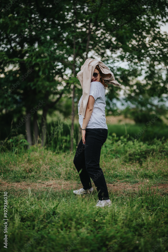 A woman playfully poses outdoors with a jacket on her head and sunglasses. She is surrounded by lush greenery, embodying a carefree and fun spirit in nature.