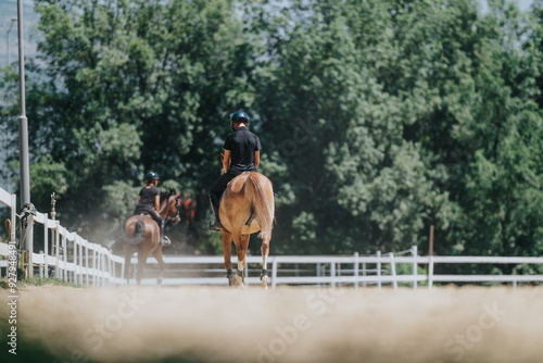 Two people are horseback riding on a dirt path with lush green trees in the background on a sunny day.