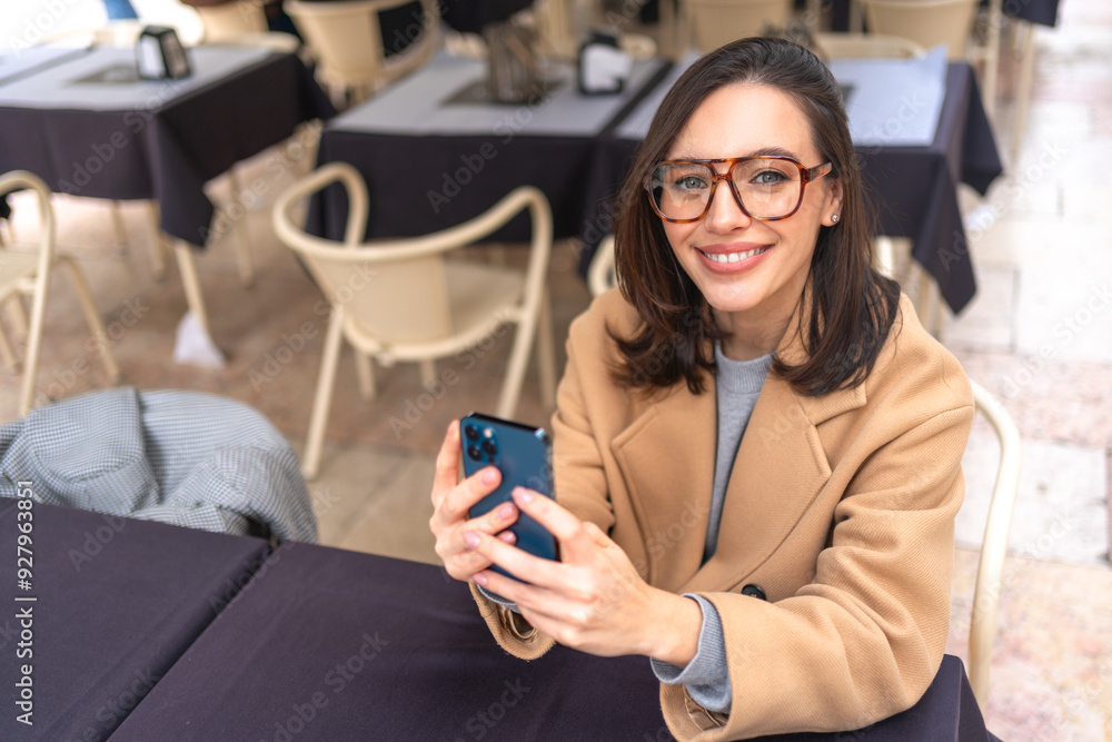 Fototapeta premium Happy middle-aged woman using smartphone sitting outdoor cafe. Smile businesswoman in glasses dressed trench coat holding smartphone in hand looking at camera. High angle view. Copy space