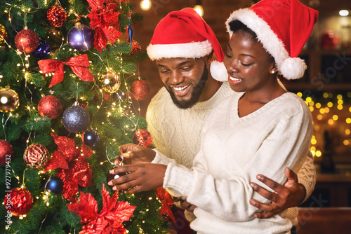 Christmas spirit. Happy black couple wearing Santa hats and decorating Xmas tree together, celebrating winter holidays at home