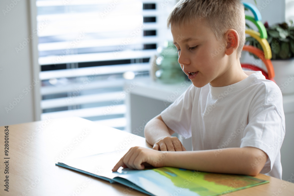 The boy is enthusiastically reading a book at a table in a bright room. Children's leisure The boy is enthusiastically reading a book at a table in a bright room. Children's leisure