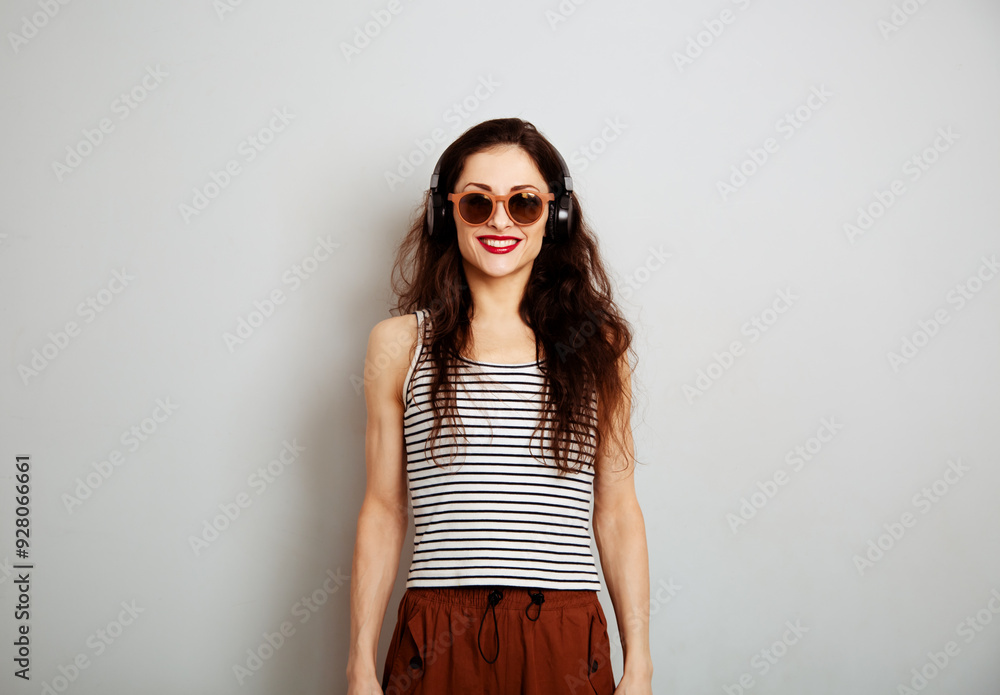 Happy toothy smiling woman in sunglasses with red lipstick listening the music in wireless headphone on white wall background with empty copy space. Lifestyle front view Happy toothy smiling woman in sunglasses with red lipstick listening the music in wireless headphone on white wall background with empty copy space. Lifestyle front view