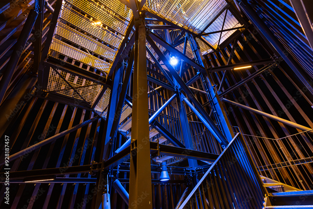 Obraz premium An upward view of a metal structure at night, dramatically lit with bright blue and yellow lights. The intricate framework and vibrant lighting create a futuristic, abstract visual effect.