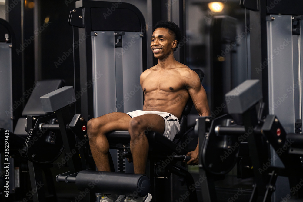 Naklejka premium Smiling shirtless strong black sportsman doing exercises on leg extensions machine.