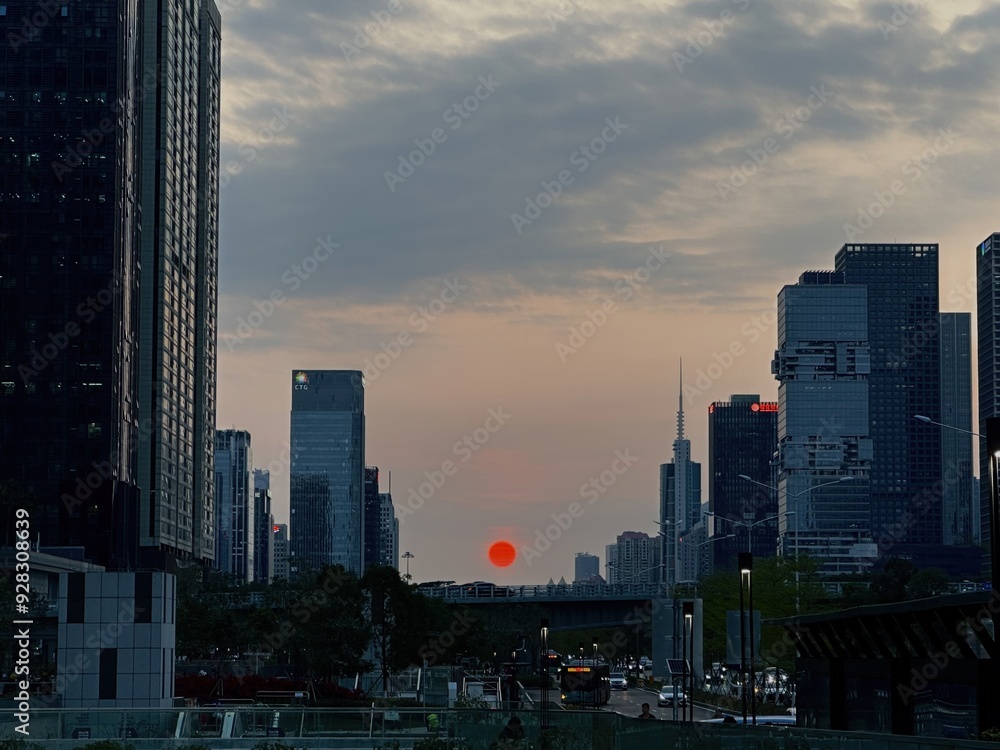 Obraz premium A vibrant red sun sets over a city skyline, casting a warm glow on the tall buildings. The clouds are a soft gray, and the city lights are beginning to twinkle.
