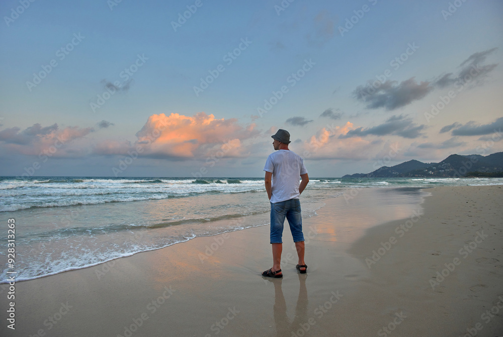 Man in white shirt, blue shorts, and hat watches sunset on sandy beach. Waves lap gently, sky is a mix of blues and pinks Man in white shirt, blue shorts, and hat watches sunset on sandy beach. Waves lap gently, sky is a mix of blues and pinks