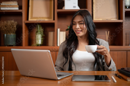 A confident, happy Asian businesswoman is having coffee while reading online papers on her laptop.