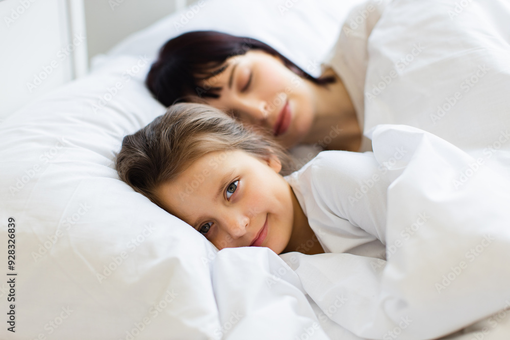 Mother and daughter lying in bed resting peacefully. Child smiling with contentment while cuddling with mother under white bed sheets. Scene highlights family bond, warmth, love, and comfort. Mother and daughter lying in bed resting peacefully. Child smiling with contentment while cuddling with mother under white bed sheets. Scene highlights family bond, warmth, love, and comfort.