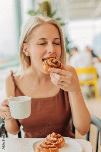 Blond woman holding coffee cup and eating cinnamon bun in cafe photo