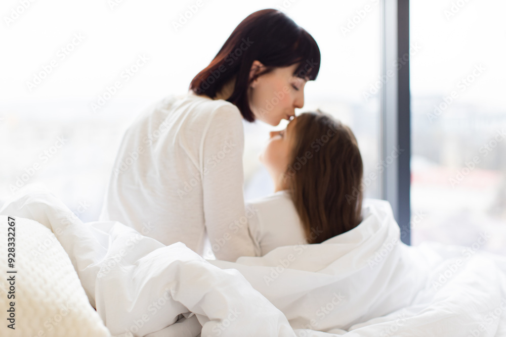 Back view. Mother kissing daughter on forehead in cozy bed by window. Warm, affectionate moment, conveying love, connection, and family bonding. White bedding and soft natural light. Back view. Mother kissing daughter on forehead in cozy bed by window. Warm, affectionate moment, conveying love, connection, and family bonding. White bedding and soft natural light.