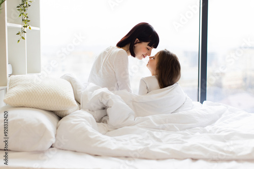 Back view of mother and daughter in bed sharing a tender moment in bright bedroom. Mother and child with noses touching, cozy white bedding. Calm, peaceful family bonding at home.