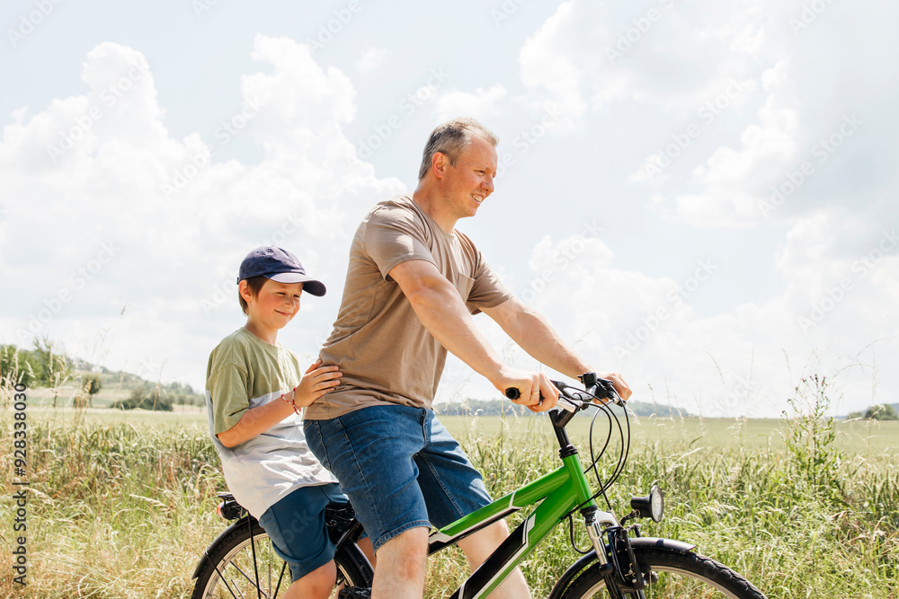 Son sitting behind father riding bicycle near field Son sitting behind father riding bicycle near field