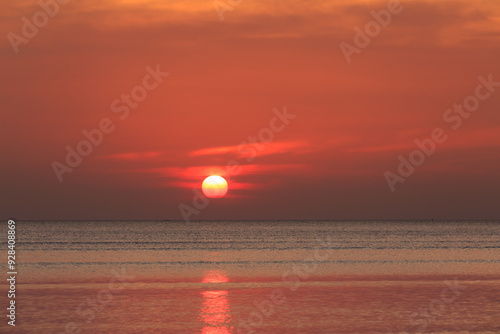 Fiery red sun dips below the horizon, painting the beach in warm hues of orange, pink, and purple. Gentle waves lap at the shore 
