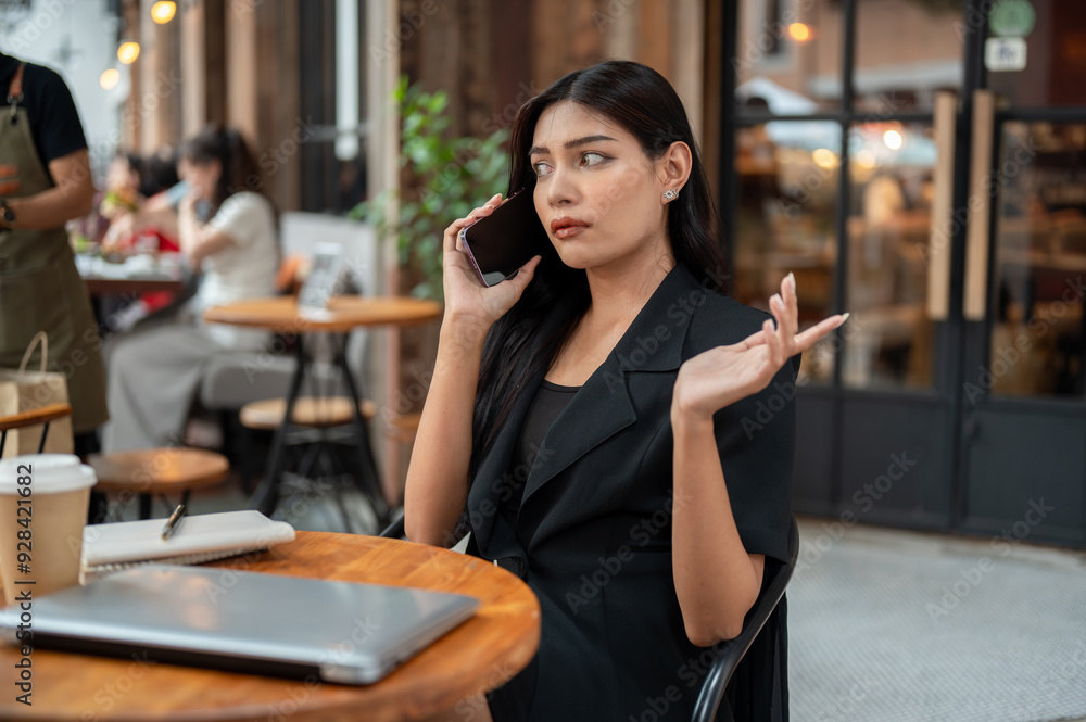 Obraz premium Uncertain, thoughtful Asian businesswoman is talking on the phone while sitting at a table of a cafe