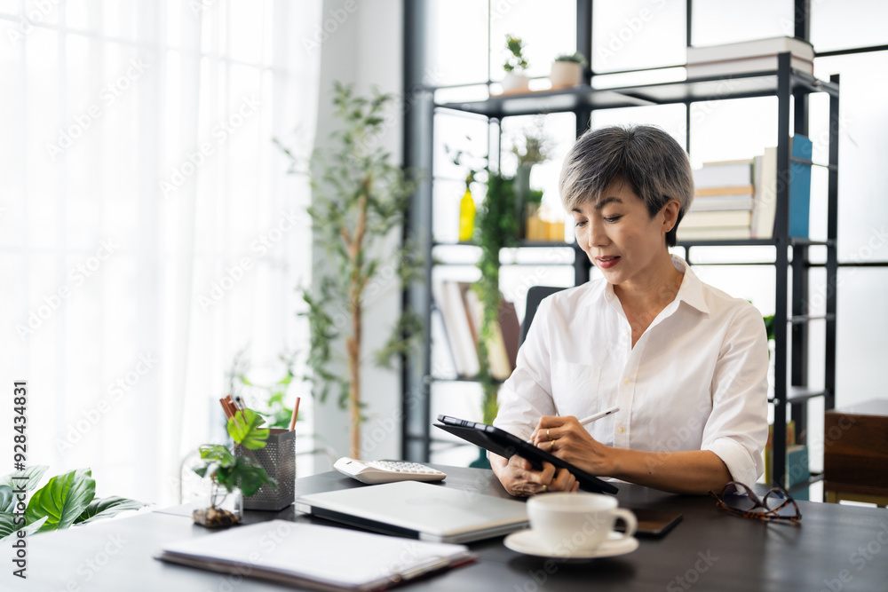 A middle-aged businesswoman is sitting at her desk in a modern office, working on a tablet with focus and concentration A middle-aged businesswoman is sitting at her desk in a modern office, working on a tablet with focus and concentration