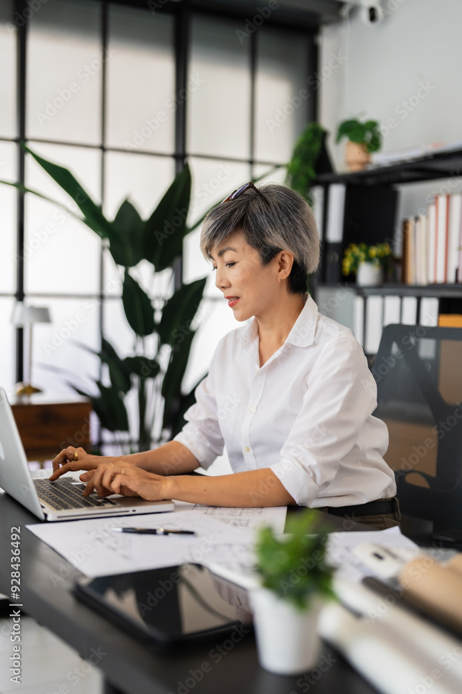 A female architect works on her laptop, surrounded by blueprints and architectural drawings, concentrating on her design projects in a tidy and modern office setting A female architect works on her laptop, surrounded by blueprints and architectural drawings, concentrating on her design projects in a tidy and modern office setting