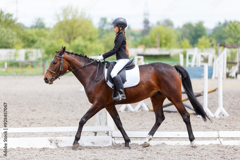 Young equestrian girl riding a chestnut horse on an outdoor arena during a cloudy day