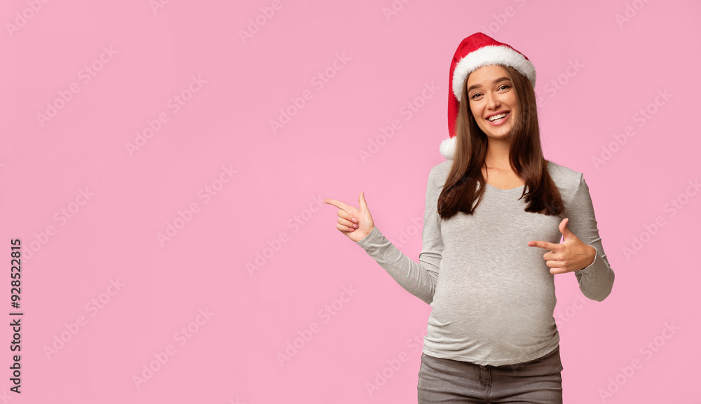Smiling Pregnant Lady In Santa Hat Pointing Fingers At Empty Space Standing In Studio On Gray Background. Pregnancy Christmas Banner Smiling Pregnant Lady In Santa Hat Pointing Fingers At Empty Space Standing In Studio On Gray Background. Pregnancy Christmas Banner